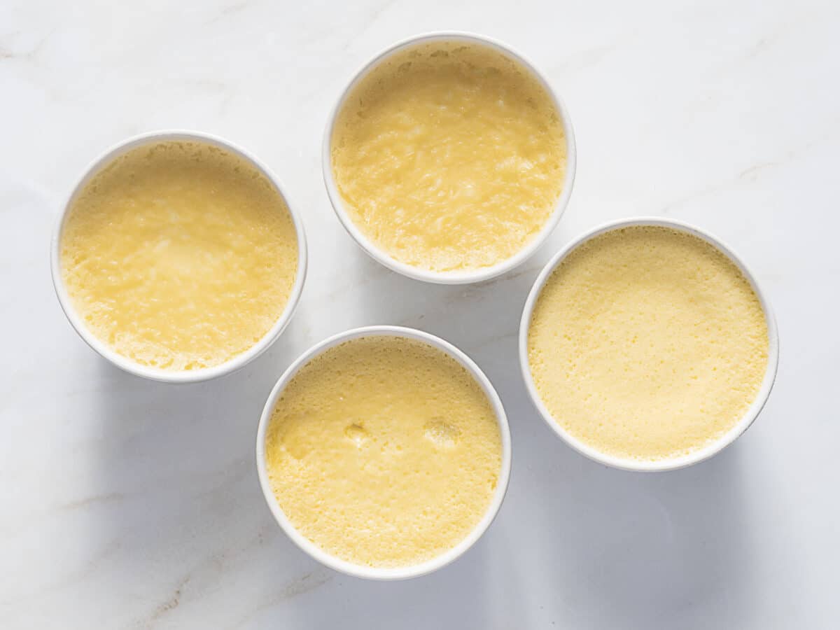 Four white ramekins filled with a smooth, yellow baked custard, arranged on a light marble surface.