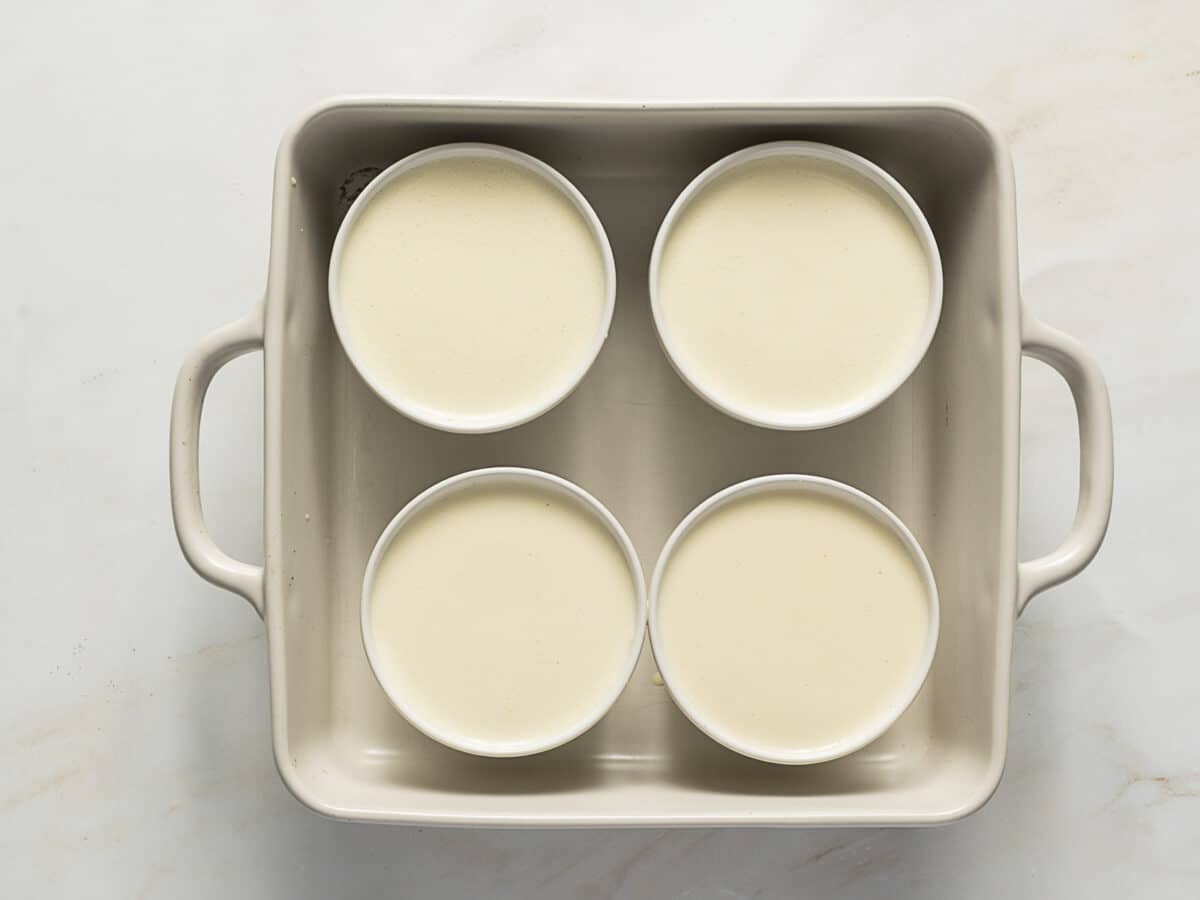 Four white ramekins filled with a pale liquid are arranged in a rectangular baking dish with handles, set on a light surface.