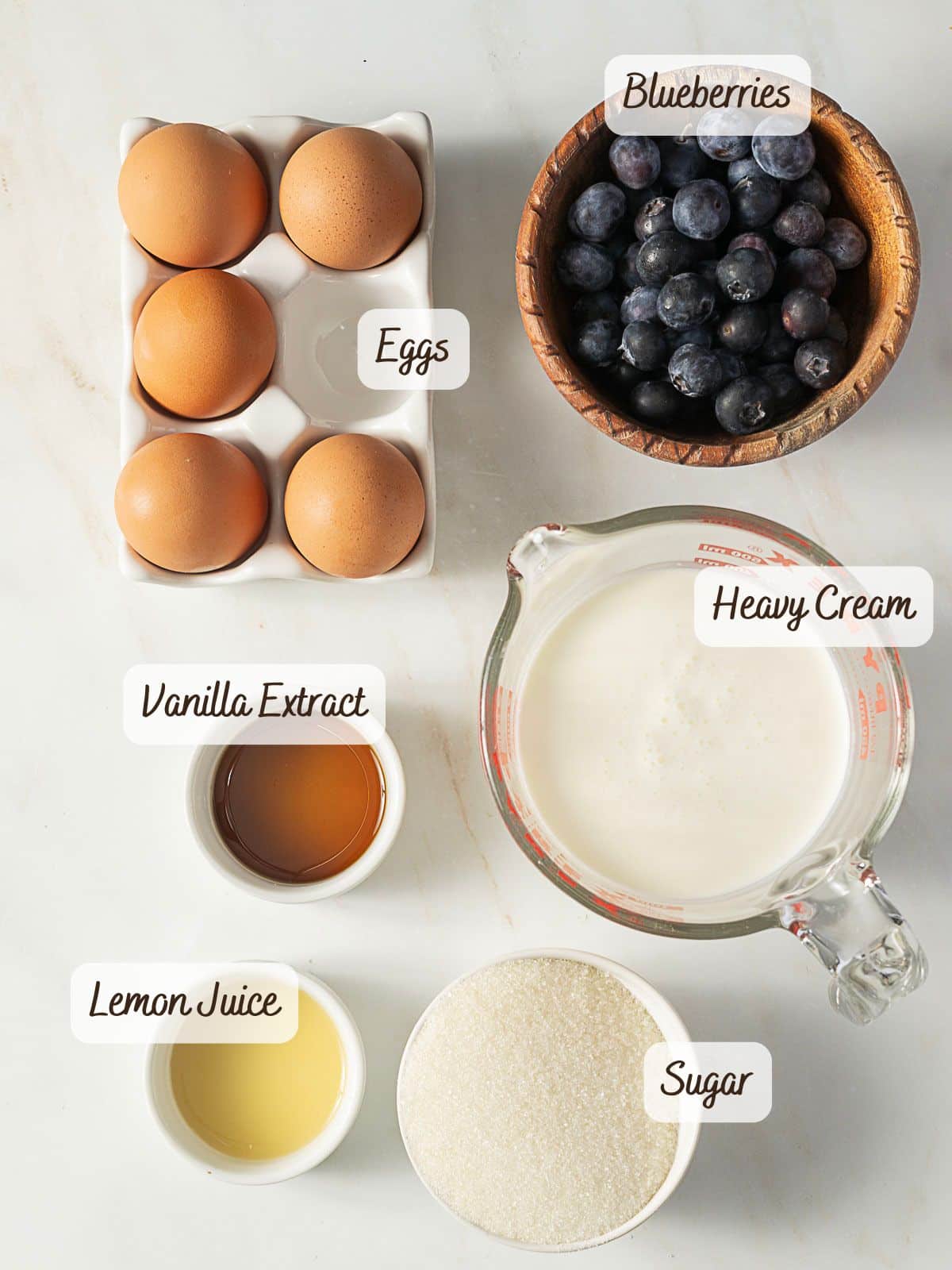 Overhead view of eggs, blueberries, heavy cream, vanilla extract, lemon juice, and sugar in bowls and containers, each labeled with its name.