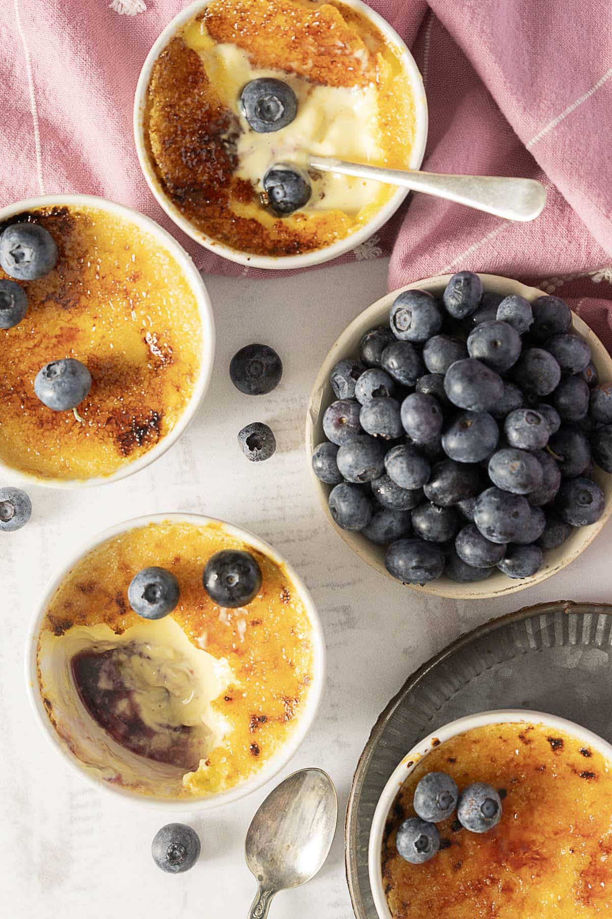 Four ramekins of crème brûlée, some topped and some garnished with blueberries, are arranged next to a bowl filled with fresh blueberries on a white surface.