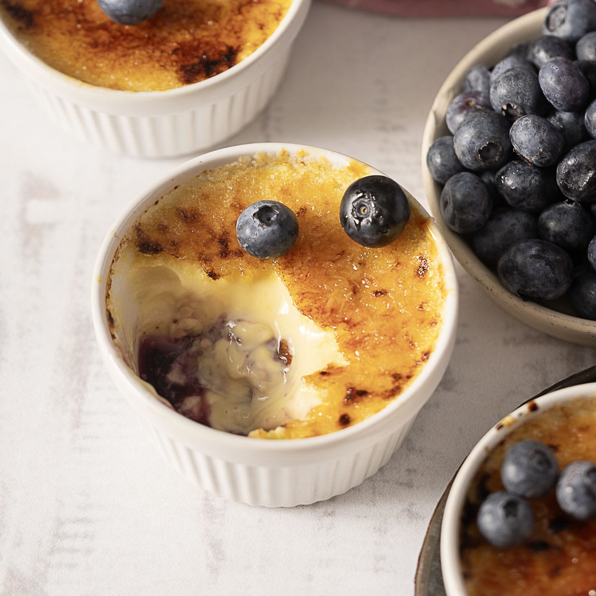 A ramekin of crème brûlée with a portion scooped out, topped with two blueberries, next to a bowl of fresh blueberries.