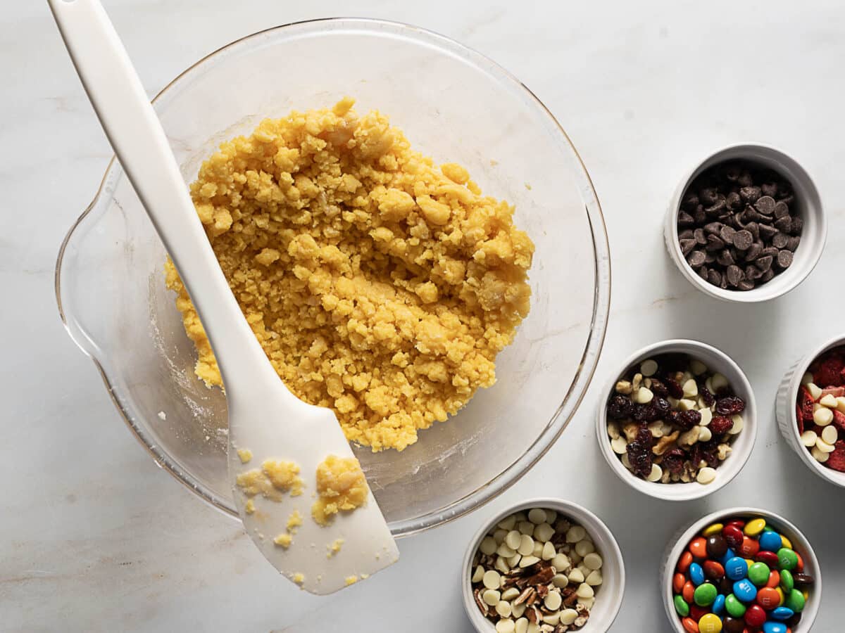A glass bowl with cookie dough and a spatula next to small bowls containing chocolate chips, white chips, candies, and dried fruit.