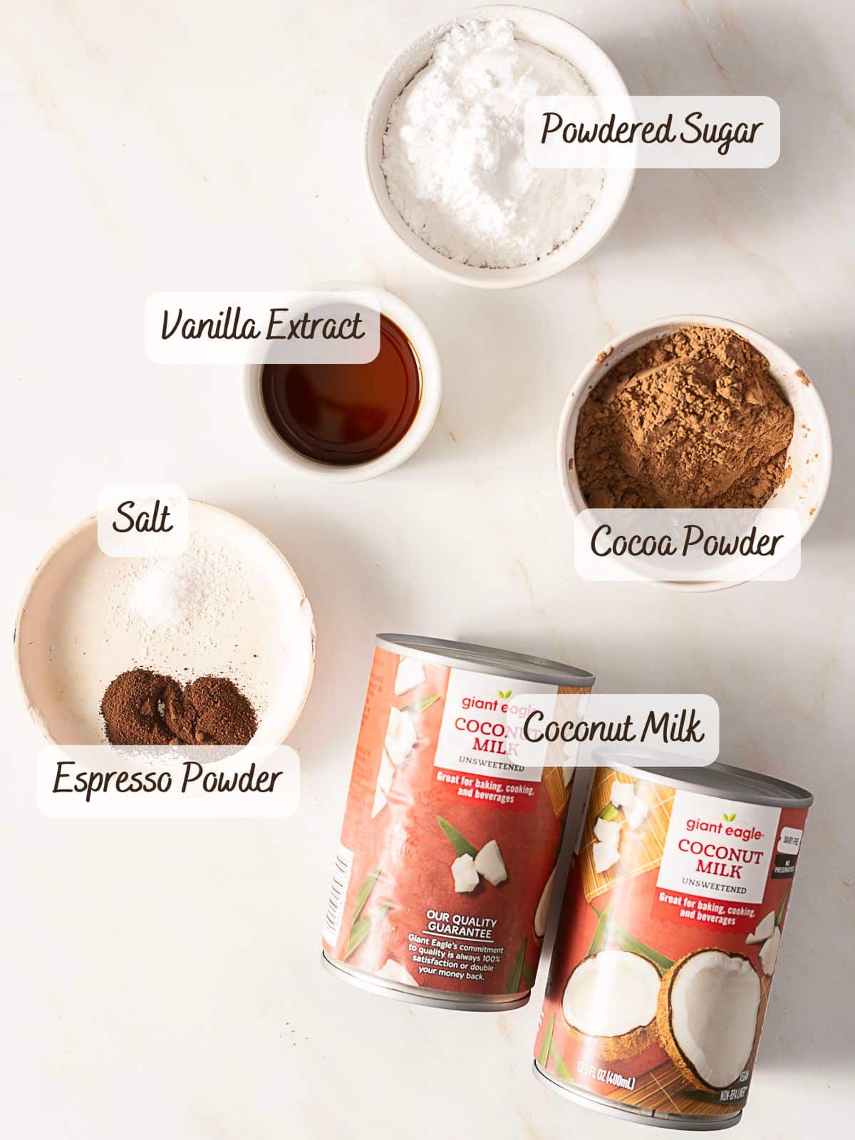 Ingredients for a dairy free chocolate mousse are arranged on a white surface: powdered sugar, vanilla extract, cocoa powder, salt, espresso powder, and two cans of coconut milk.