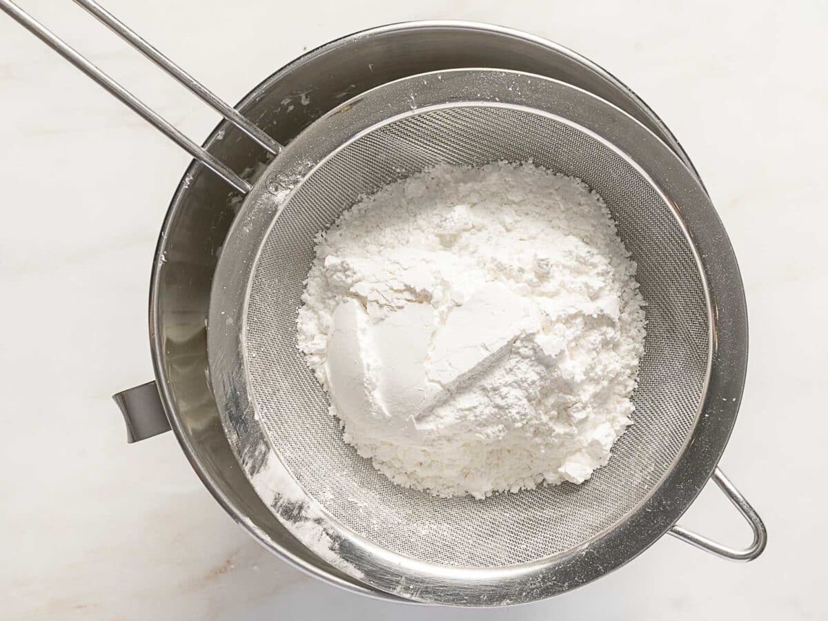 A metal sifter filled with white flour rests on top of a metal mixing bowl, viewed from above on a light surface.