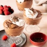 Three glass dishes of chocolate mousse topped with whipped cream and chocolate shavings, with a bowl of mixed berries and a small bowl of chocolate shavings nearby.