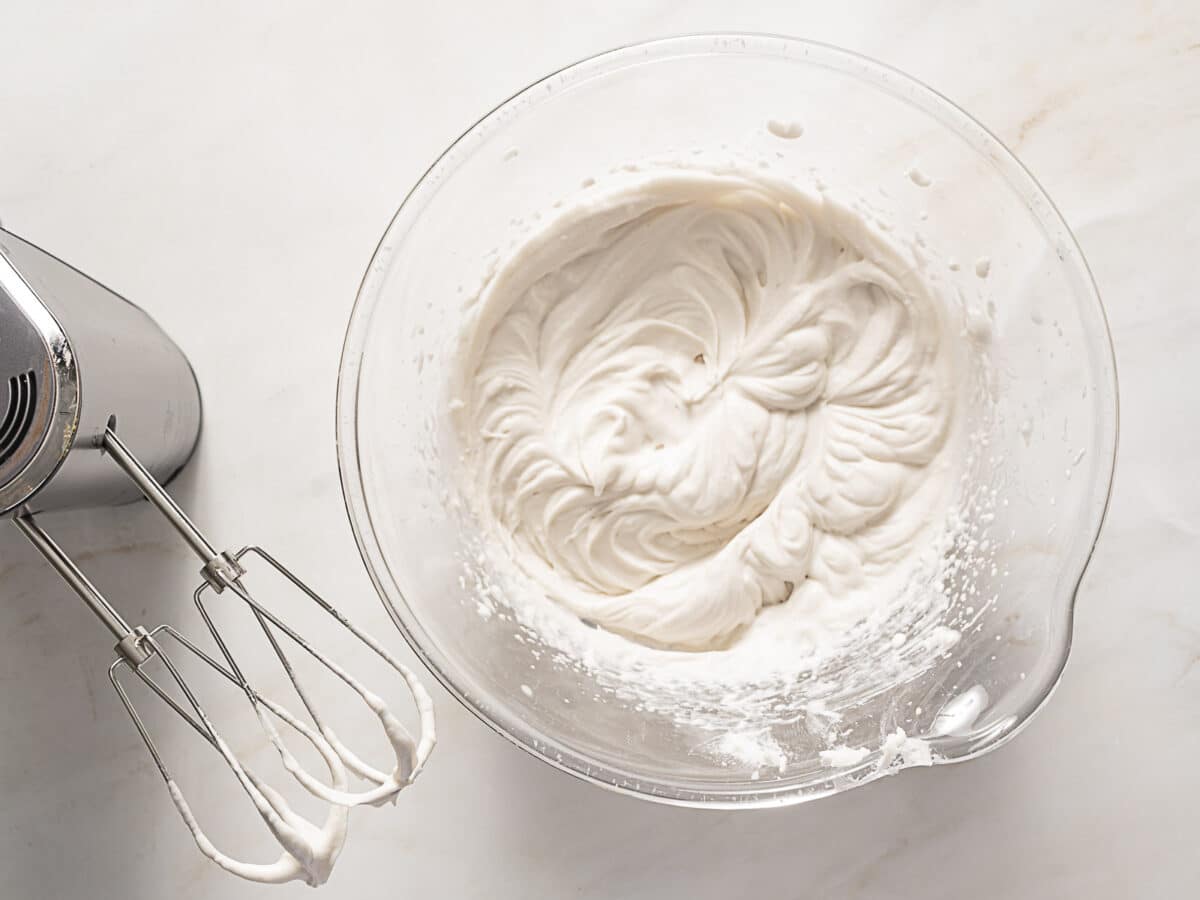 A glass bowl filled with whipped cream sits on a white surface next to an electric hand mixer with beaters covered in cream.