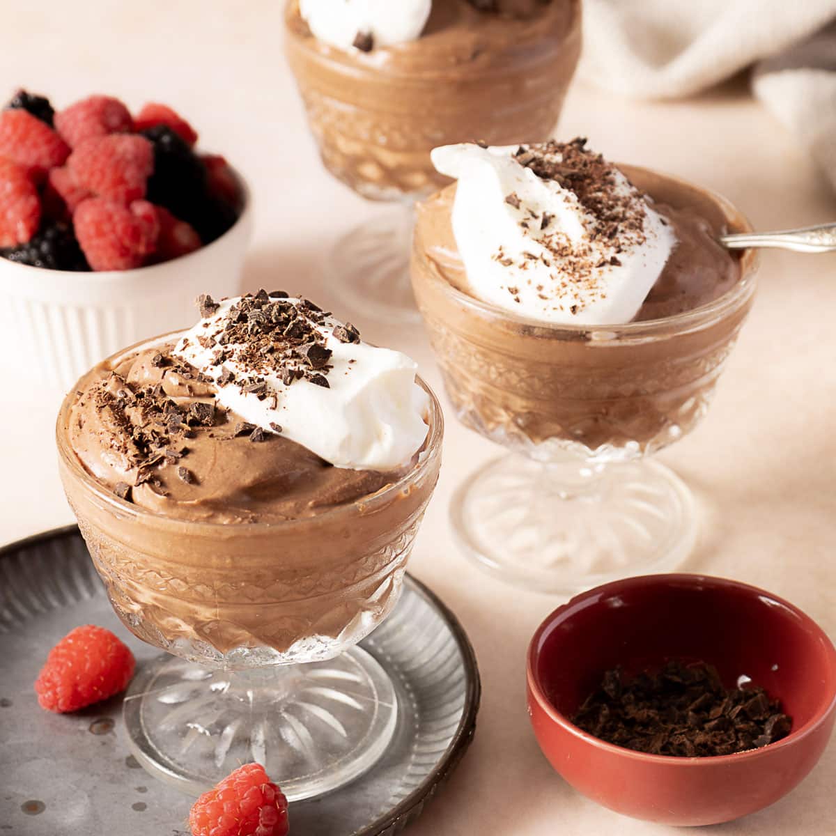Three glass dishes of chocolate mousse topped with whipped cream and chocolate shavings, with a bowl of mixed berries and a small bowl of chocolate shavings nearby.