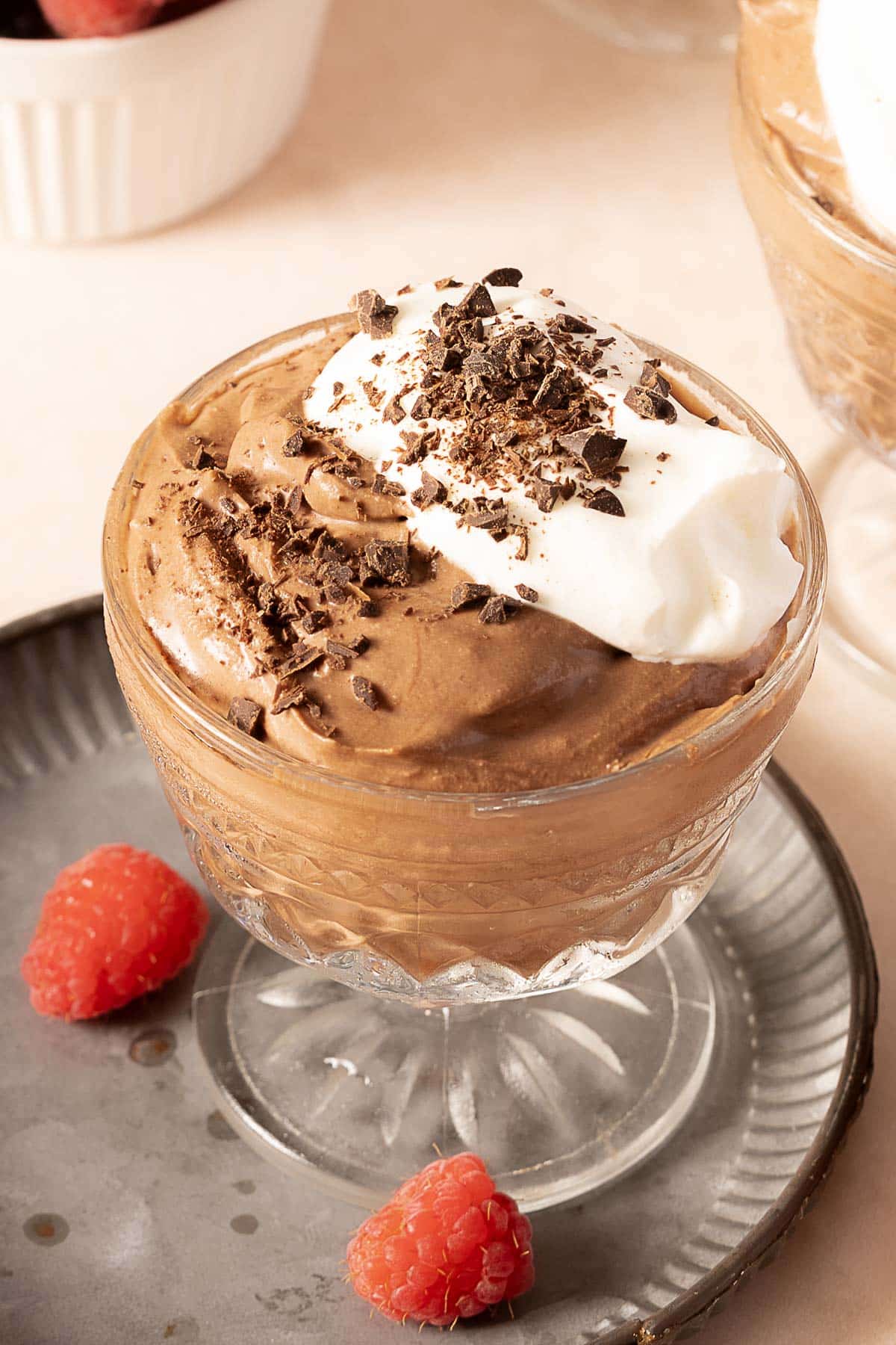 A glass dish of chocolate mousse topped with whipped cream and chocolate shavings, served with two raspberries on a gray plate.