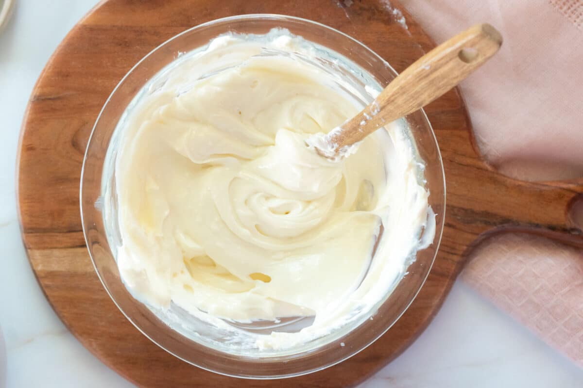 A glass bowl filled with creamy white frosting and a wooden spoon, placed on a round wooden cutting board.