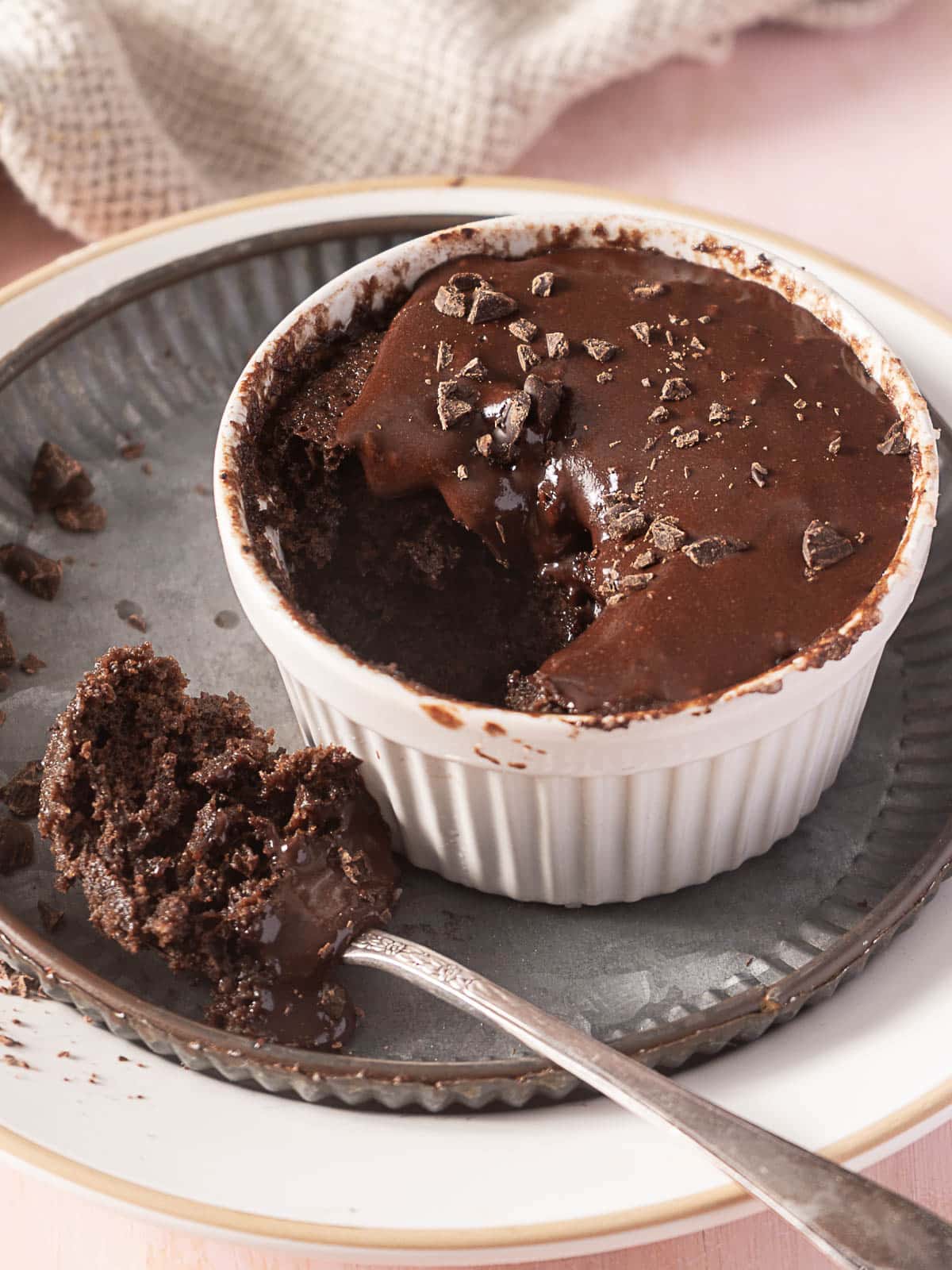 A chocolate lava cake in a white ramekin with a spoonful removed, showing a molten chocolate center, on a gray plate with scattered chocolate pieces.
