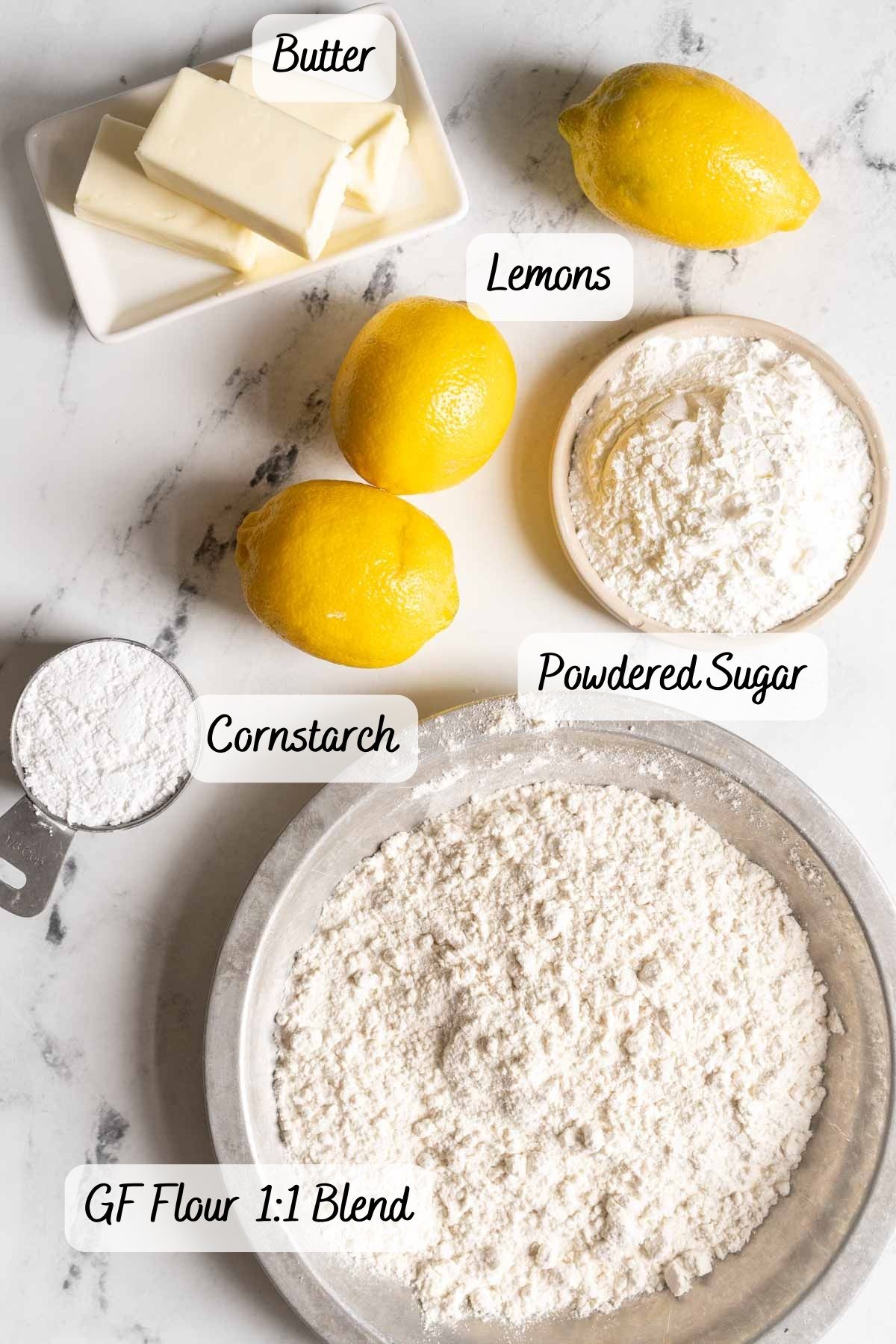 Overhead view of labeled baking ingredients on a marble counter: butter, lemons, powdered sugar, cornstarch, and a bowl of gluten-free 1:1 flour blend.