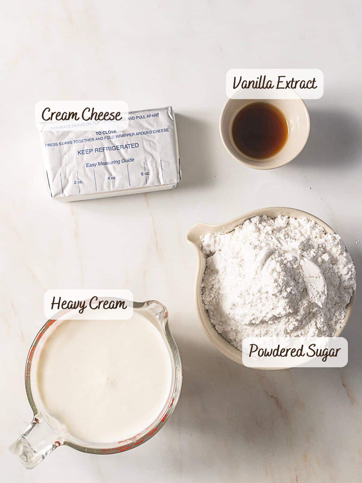 Four labeled baking ingredients on a white surface: a block of cream cheese, a bowl of vanilla extract, a measuring cup of heavy cream, and a bowl of powdered sugar—perfect for making cream cheese frosting without butter.