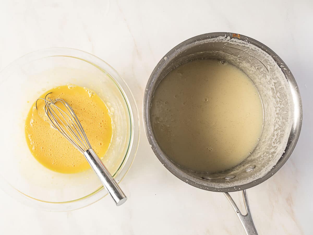 A glass bowl with whisked yellow egg mixture and a whisk sits next to a saucepan containing a thick, pale liquid on a marble surface.
