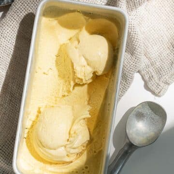 Rectangular container of yellow ice cream with two scoops removed, an ice cream scooper beside it, and a textured cloth in the background.
