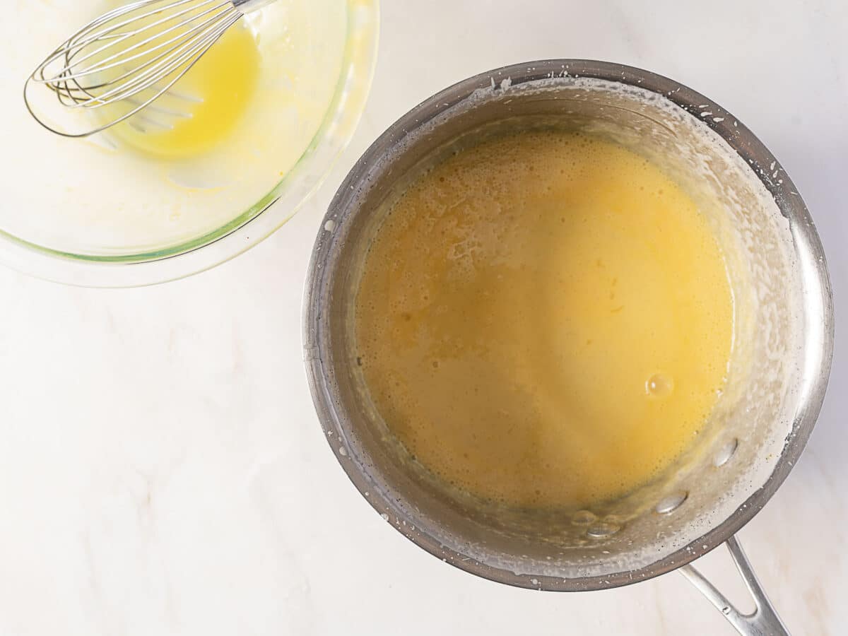 A saucepan with yellow custard mixture on a white surface, next to a glass bowl containing remnants of the same mixture and a metal whisk.