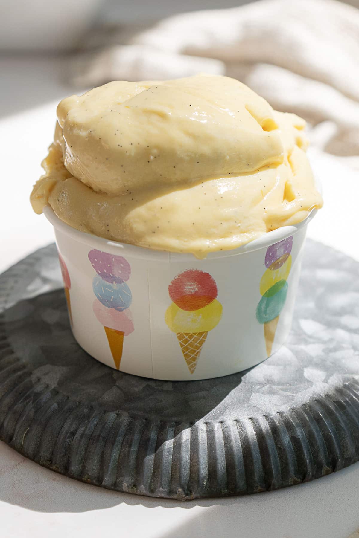 A paper cup with colorful ice cream cone designs is filled with scoops of yellow ice cream, placed on a round metal tray.