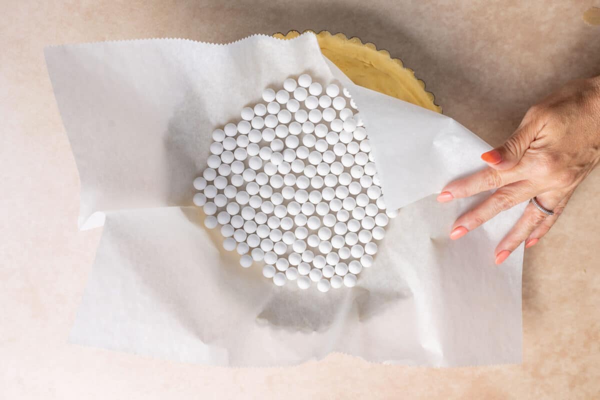A hand holds baking parchment filled with ceramic pie weights over an unbaked pie crust in a tart pan.