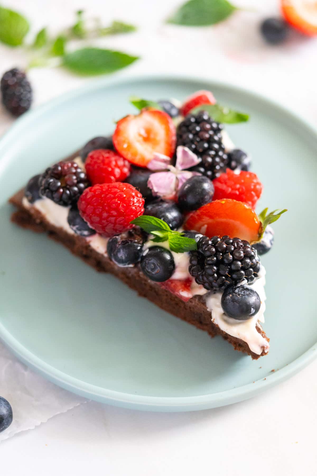 A slice of dessert pizza topped with cream, strawberries, blackberries, raspberries, blueberries, and mint leaves on a light blue plate.