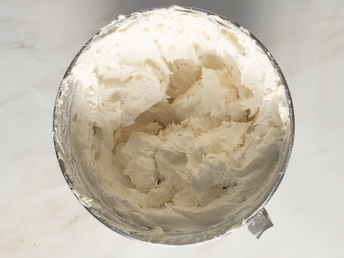 A metal mixing bowl filled with creamy, whipped buttercream frosting on a light-colored surface.