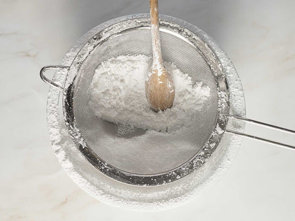 A metal mesh sieve with powdered sugar and a wooden spoon sits on a white surface, with some sugar dusted around the sieve.