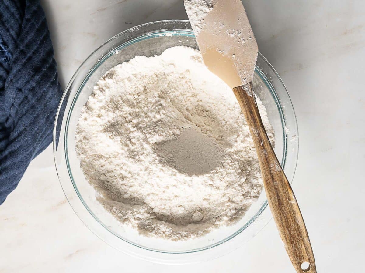 A glass bowl filled with flour and yeast, with a wooden-handled spatula resting on the side, placed on a white surface.