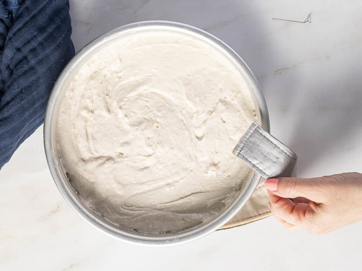 A hand holding a round metal cake pan filled with smooth, swirled gluten free buttermilk cake batter on a marble surface.