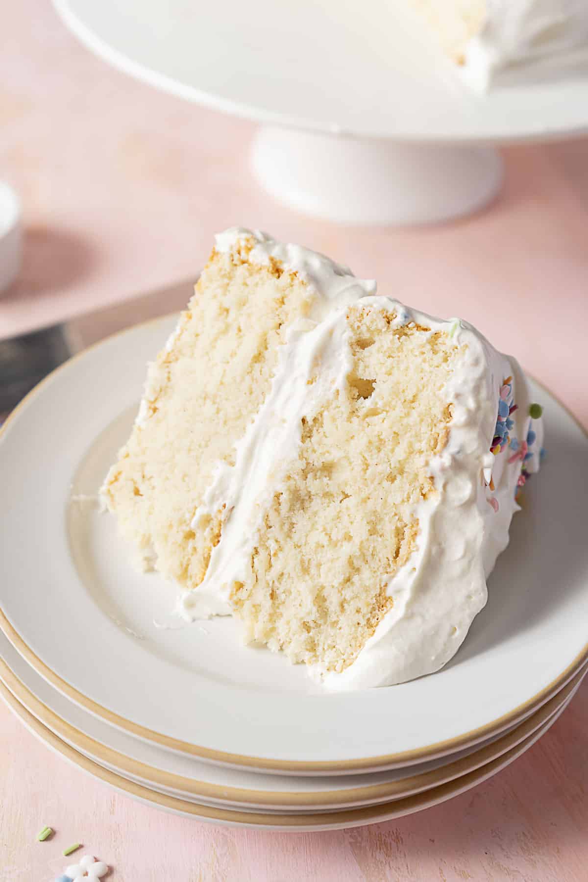 A slice of white layer cake with white frosting on a stack of white plates, with the rest of the cake in the background.