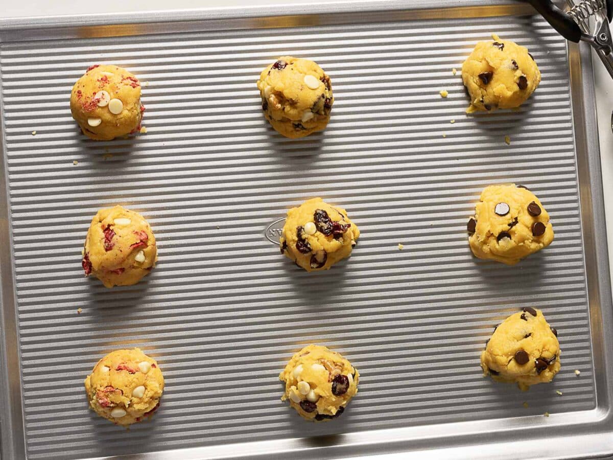 Nine unbaked cookie dough balls with chocolate and white chips are arranged in rows on a ribbed metal baking sheet.