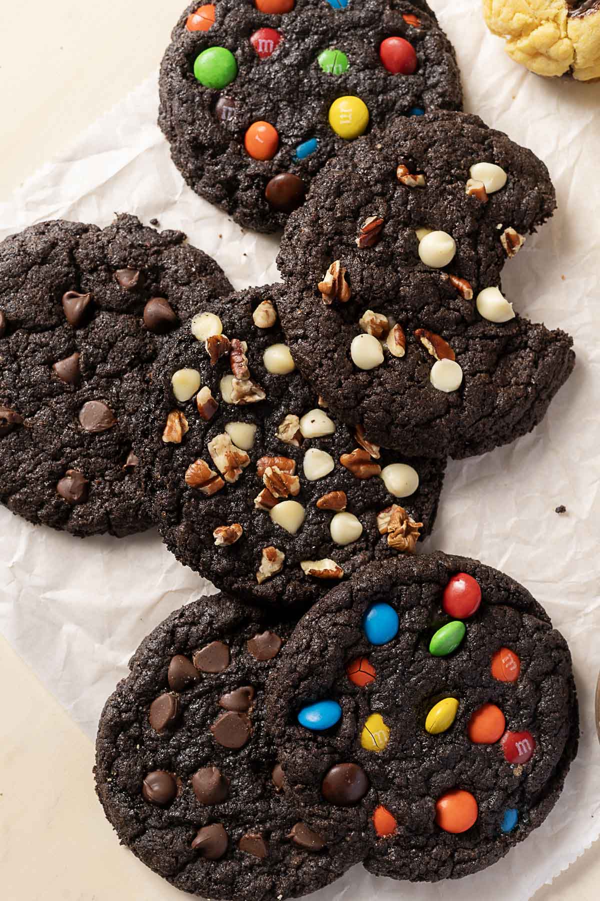 Six large, dark chocolate cookies on parchment paper, topped with assorted mix-ins including colorful candies, white chips, pecans, and chocolate chips.