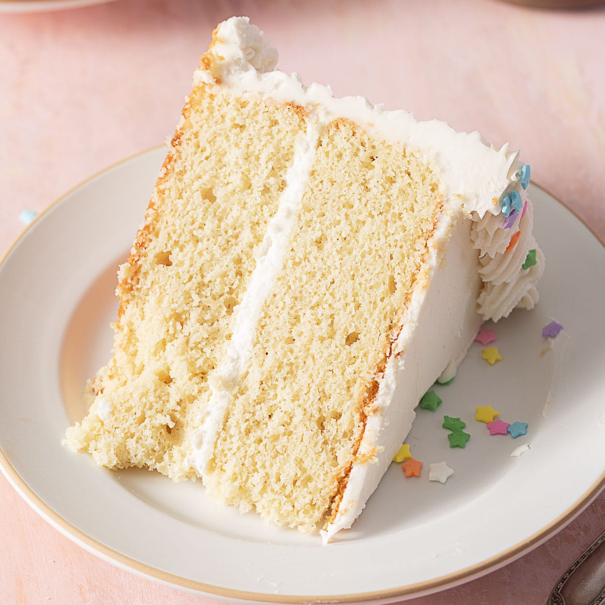 A slice of two-layer vanilla cake with white frosting on a plate, decorated with colorful star-shaped sprinkles.