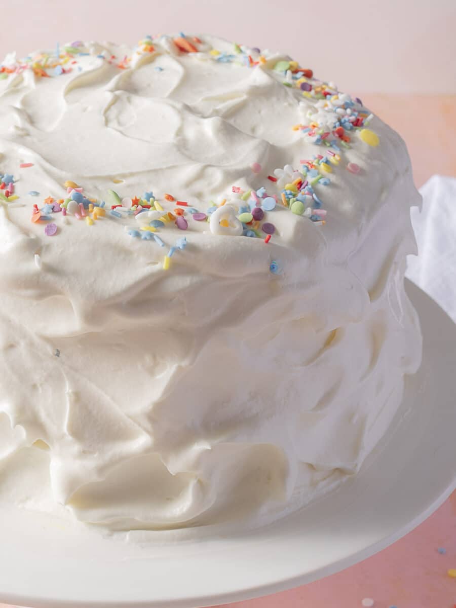 A frosted layer cake on a white stand, decorated with pastel-colored sprinkles around the top edge.