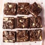 Nine cheesecake swirl brownies are arranged in a 3x3 grid on parchment paper, showing marbled patterns of dark chocolate and cream cheese.