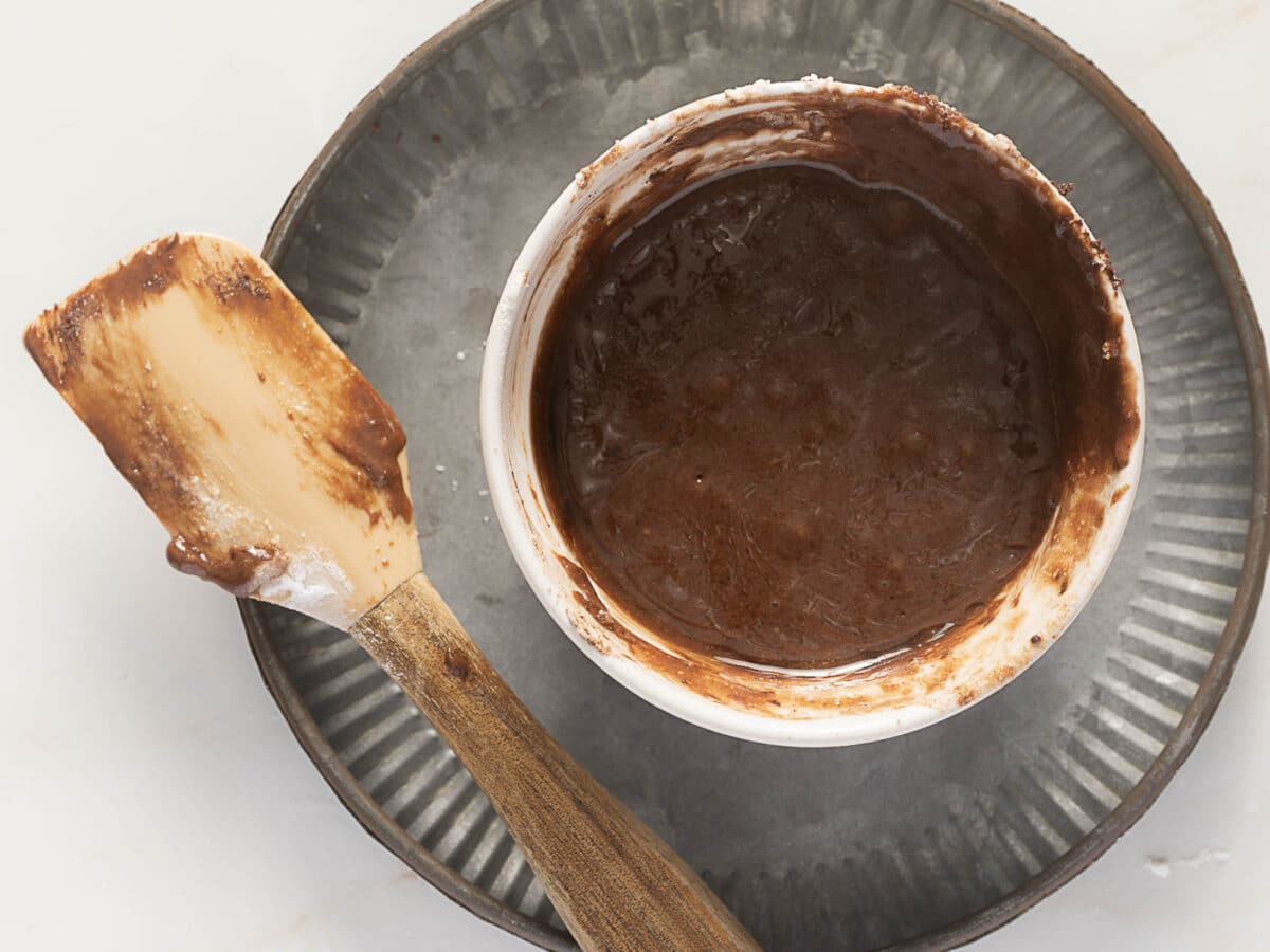 A bowl of chocolate cake batter sits on a round metal tray next to a used spatula with batter residue.