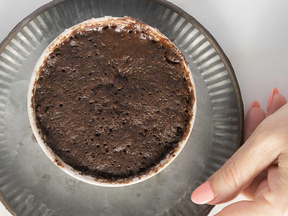A hand holds a metal plate with a cup containing a baked chocolate mug cake viewed from above.