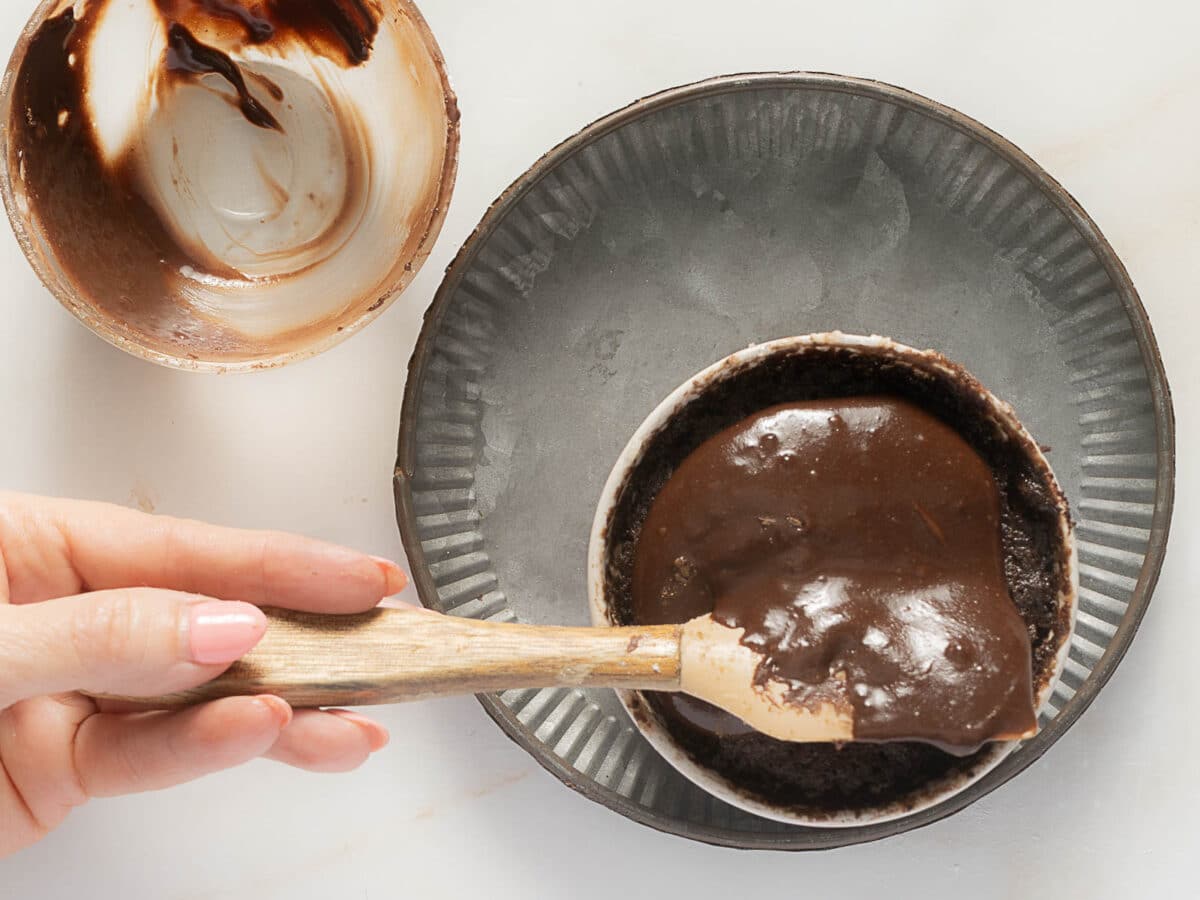 A hand uses a spatula to spread chocolate batter in a round baking dish, with an empty mixing bowl nearby on a white surface.