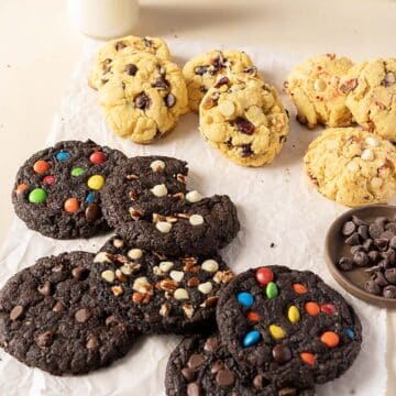 Assorted cookies with chocolate chips, white chocolate chips, pecans, and colorful candy-coated chocolates on parchment paper, with a small bowl of chocolate chips nearby.
