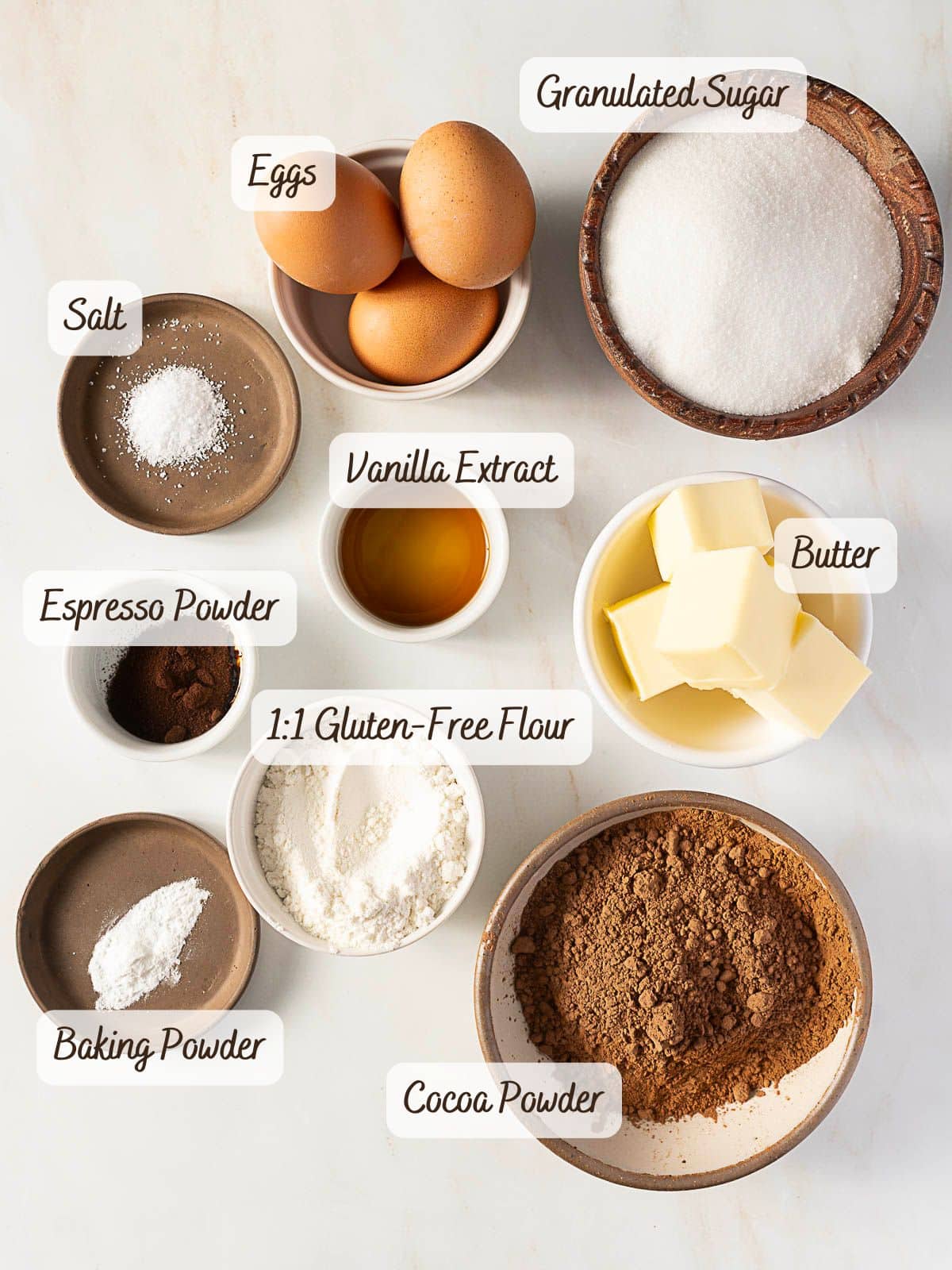 Overhead view of labeled baking ingredients in bowls for a gluten free dessert pizza: eggs, granulated sugar, salt, vanilla extract, butter, espresso powder, gluten-free flour, cocoa powder, and baking powder.