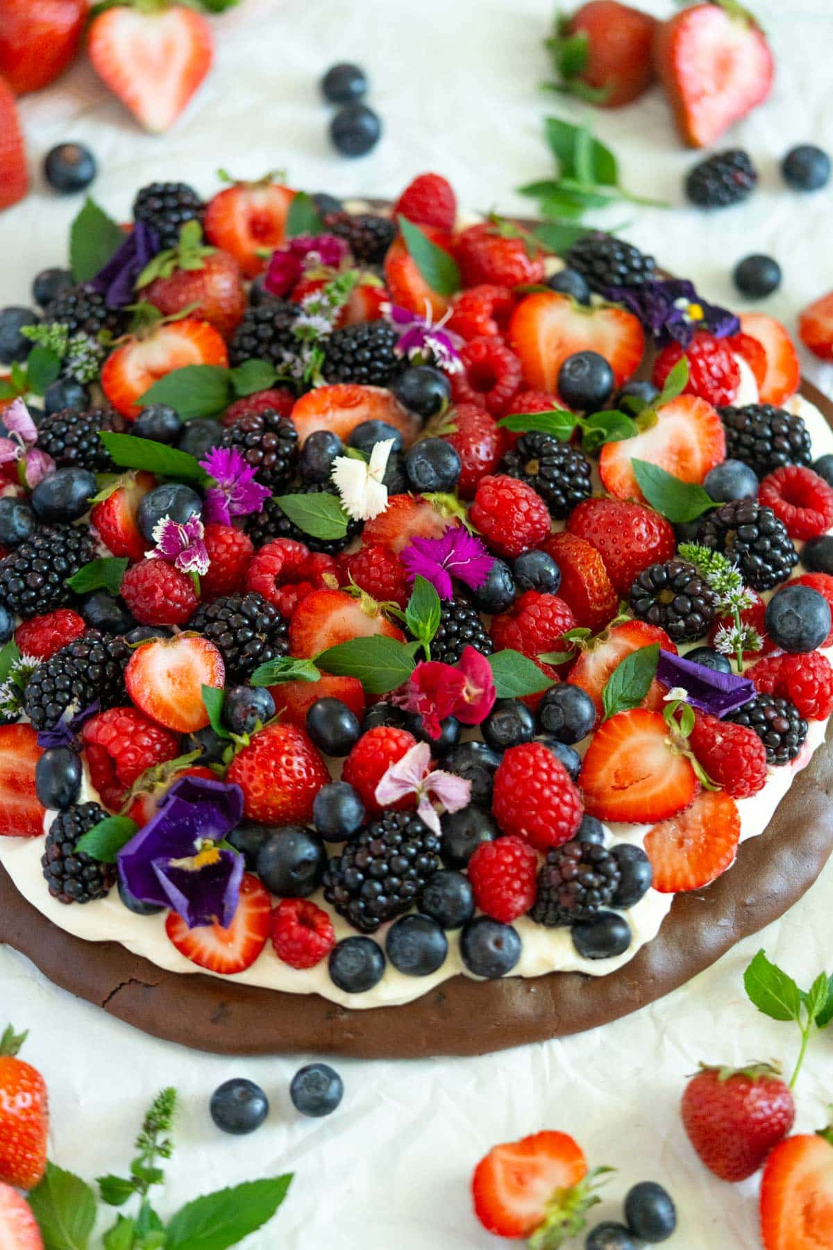 A dessert topped with assorted fresh berries, edible flowers, and green leaves, set on a layer of cream and a chocolate base, surrounded by scattered berries and mint.