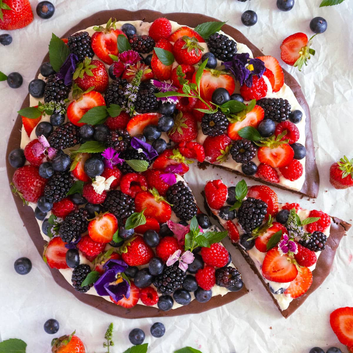 A fruit pizza topped with cream, strawberries, blackberries, blueberries, and edible flowers, with one slice partially removed, on a white surface.