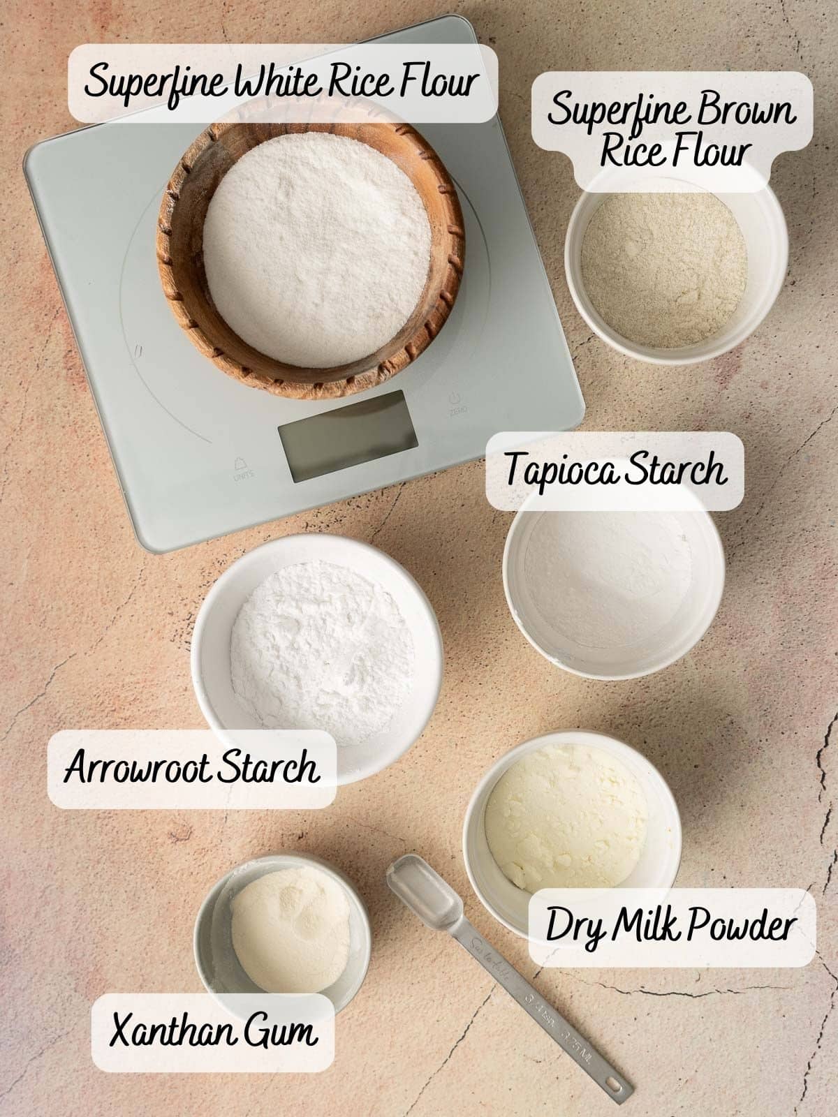 Seven white bowls on a beige surface hold labeled baking ingredients: superfine white and brown rice flour, tapioca starch, arrowroot starch, dry milk powder, and xanthan gum. A scale is visible.