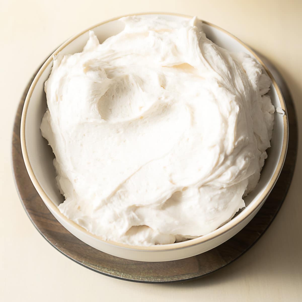 A white ceramic bowl filled with thick, fluffy gluten free frosting sits on a wooden coaster against a light background.