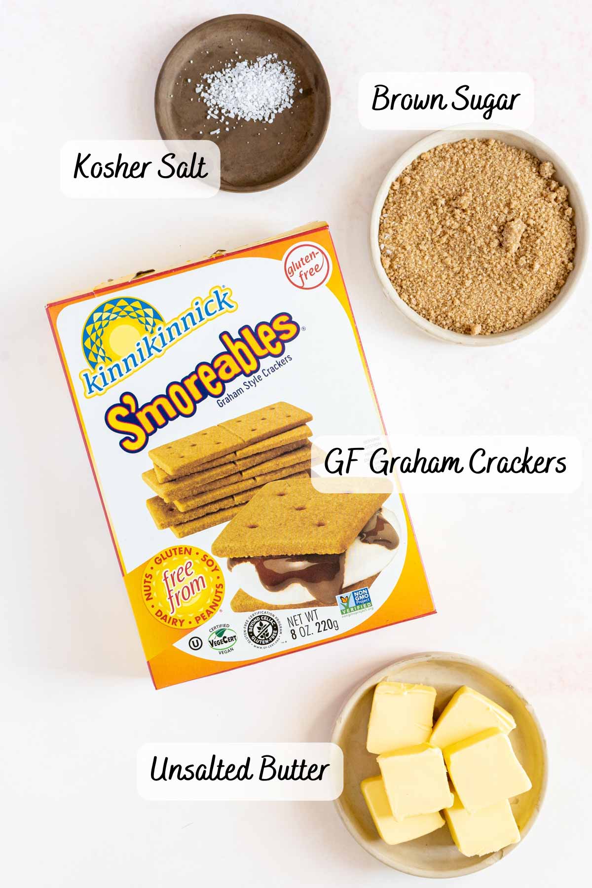 A box of gluten-free graham crackers, a bowl of brown sugar, a small dish of kosher salt, and a plate of unsalted butter are arranged on a white surface.
