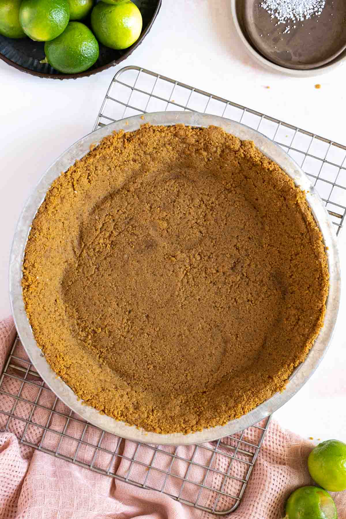 A prepared graham cracker pie crust sits in a round metal pie dish on a cooling rack, surrounded by whole limes and kitchen towels.