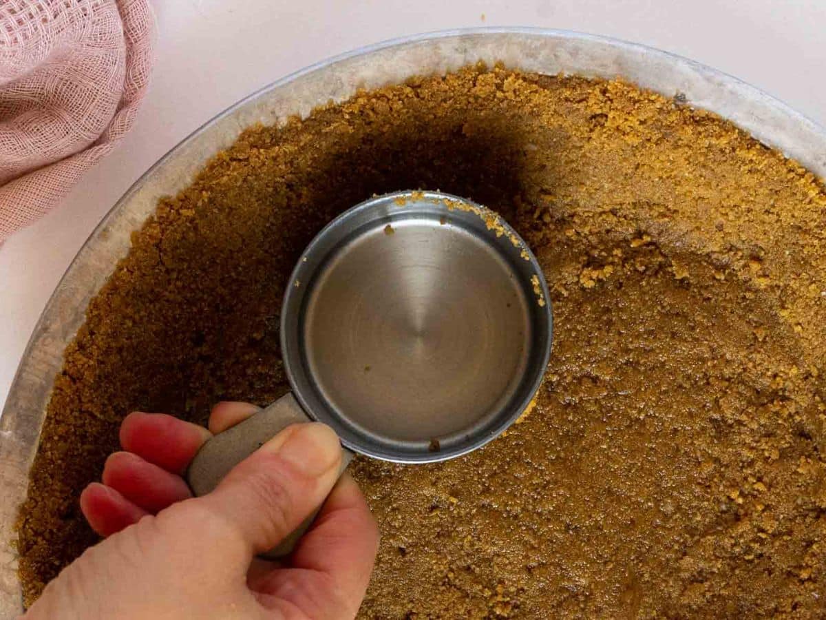 A hand presses a metal measuring cup onto a graham cracker pie crust in a pie dish.