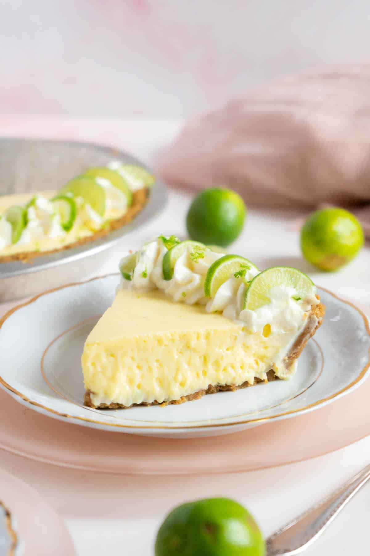 A slice of key lime pie topped with whipped cream and lime slices sits on a plate, with whole limes and the rest of the pie in the background.