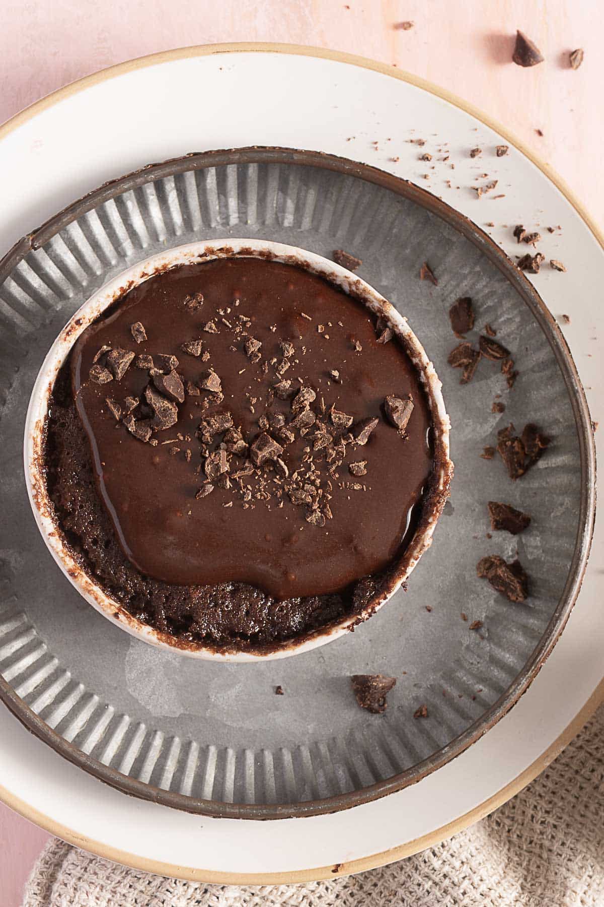 A chocolate mug cake topped with melted chocolate and chocolate shavings sits on a gray plate with crumbs scattered around.
