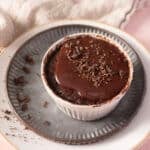 A chocolate lava cake in a white ramekin, topped with chocolate sauce and chocolate shavings, sits on a plate with crumbs around it.
