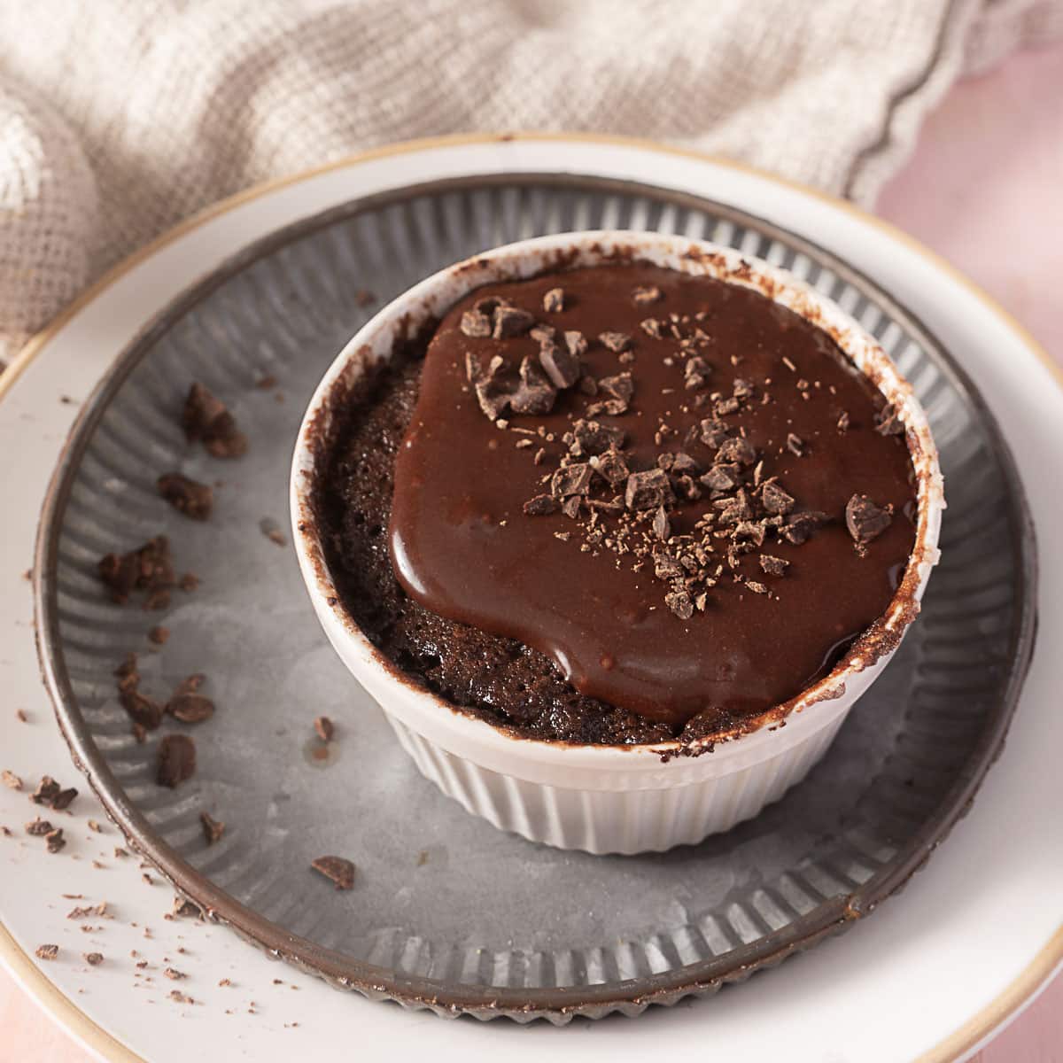 A chocolate lava cake in a white ramekin, topped with chocolate sauce and chocolate shavings, sits on a plate with crumbs around it.