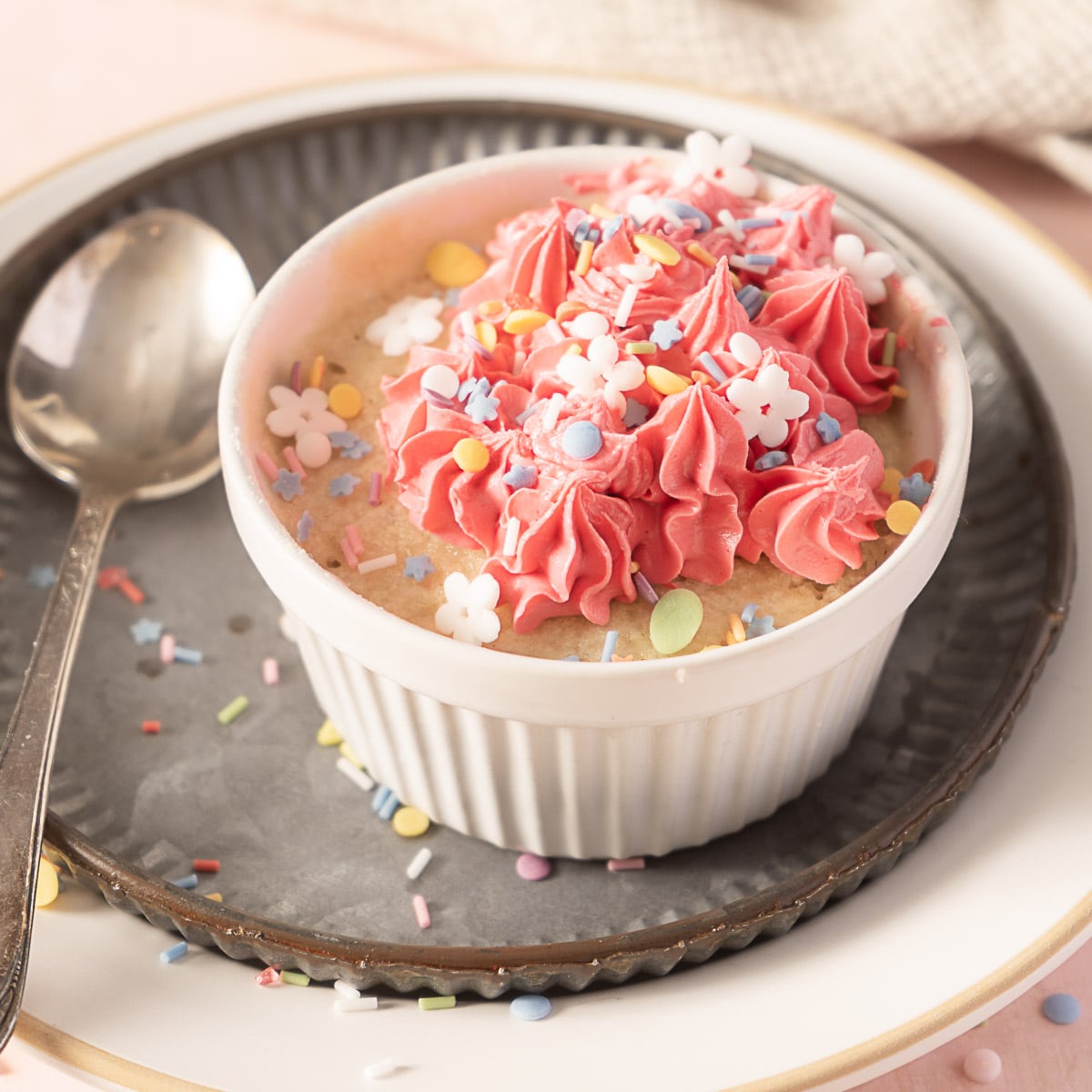A small white ramekin filled with cake, topped with pink frosting and colorful flower-shaped sprinkles, sits on a metal plate with a spoon beside it.