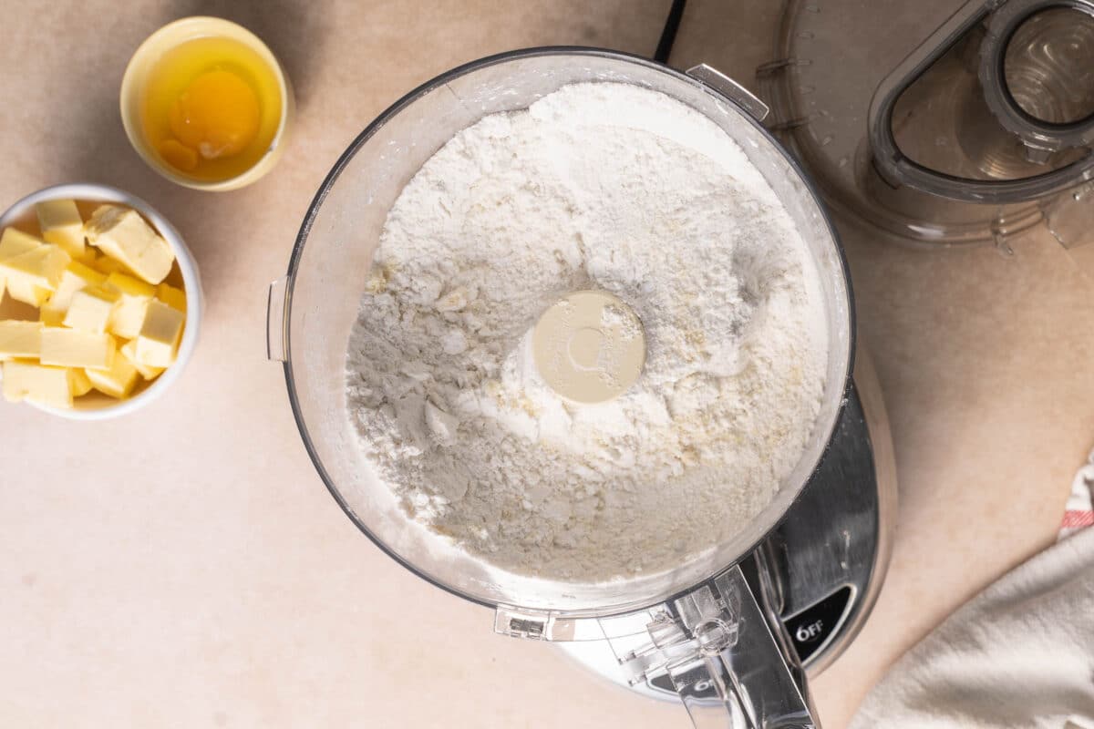 A food processor bowl filled with flour sits on a counter next to a bowl with butter cubes and a bowl containing an egg and egg yolk.
